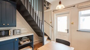 A compact kitchen with a built-in microwave, toaster, and shelves. A wooden table and chairs are positioned near a white door with a window. - Host & Stay | The Railman's House (York)