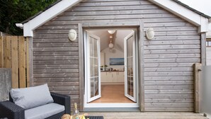 A wooden cottage with a balcony, a wicker sofa, and a table with fruits and a bottle. - Kerensa is a cosy and detached barn conversion located in the lovely seaside village of Portreath (Portreath)