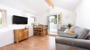 A modern living room with a grey sofa, wooden dining table, and a TV mounted on the wall. - Kerensa is a cosy and detached barn conversion located in the lovely seaside village of Portreath (Portreath)