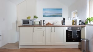 A modern kitchen with white cabinets, a microwave, a kettle, and a coffee maker. There is a painting on the wall and a plant on the counter. - Kerensa is a cosy and detached barn conversion located in the lovely seaside village of Portreath (Portreath)