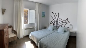 A bedroom with two beds, a wooden dresser, a wicker chair, a radiator, and a wall-mounted artwork.