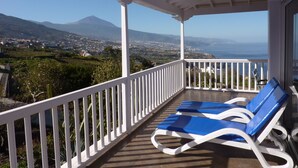 A balcony with two blue lounge chairs, a white railing, and a view of a coastal landscape with mountains in the distance.