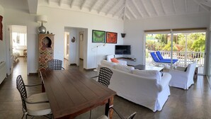 A spacious living area with a wooden dining table, white sofas, and a balcony with blue lounge chairs.