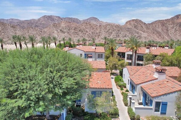 A residential area with multiple houses featuring red-tiled roofs, surrounded by greenery and palm trees, with a mountainous backdrop.