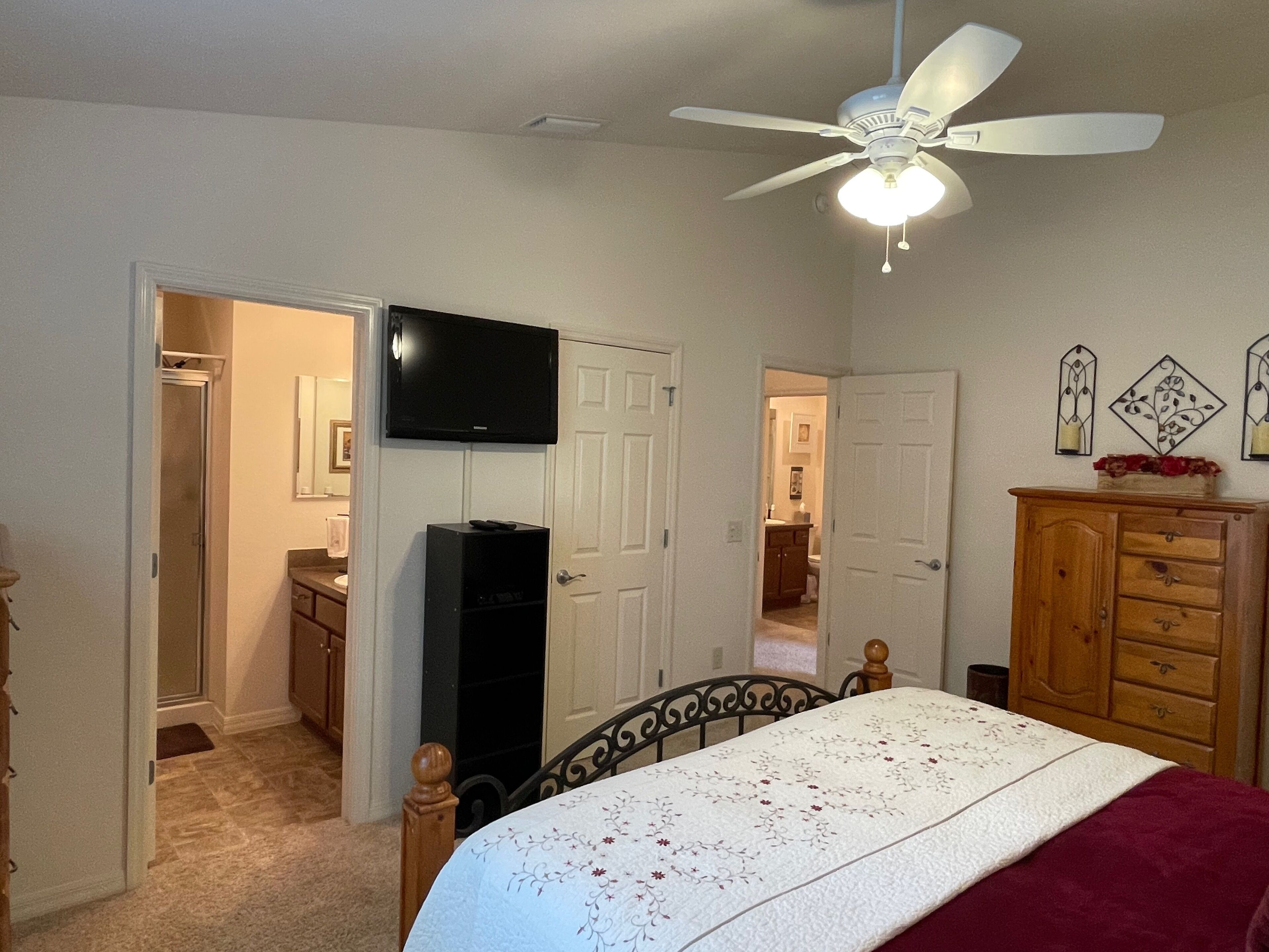 A bedroom with a bed, a ceiling fan, a TV, a wooden dresser, and a door leading to another room.
