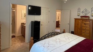 Un dormitorio con cama, ventilador de techo, televisión, cómoda de madera y puerta que da a otra habitación.