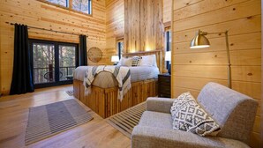 Interior d'una cabana de fusta amb un llit, un sofà, una làmpada de peu i una finestra amb vista al bosc.