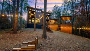 Una cabana de fusta moderna amb un gran terrat i llums lluminoses, envoltada d'arbres.