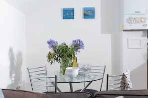 Uma sala de jantar com mesa de vidro, cadeiras, um vaso com flores e uma parede branca com duas molduras.