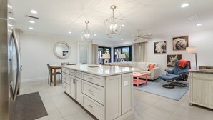 A modern kitchen with a central island, a dining area with a round table, and a living space with a sofa and armchairs. - Waters Edge 104s 2 Bedroom Condo by RedAwning (Holmes Beach)