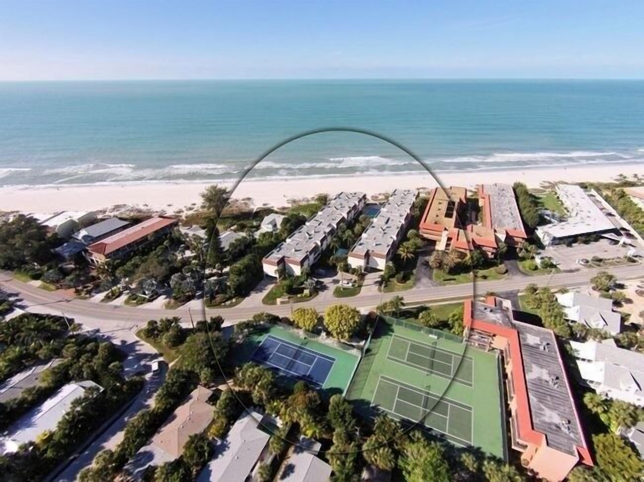 An aerial view of a coastal residential area with a tennis court, beach, and ocean.