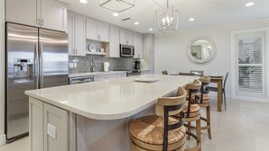 A modern kitchen with a large island, stainless steel appliances, and a dining area with wooden chairs. - Waters Edge 104s 2 Bedroom Condo by RedAwning (Holmes Beach)