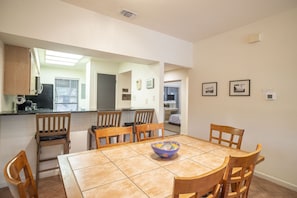A dining area with a tiled table, wooden chairs, and a bowl. There is a kitchen area with a microwave and cabinets visible through an open doorway.