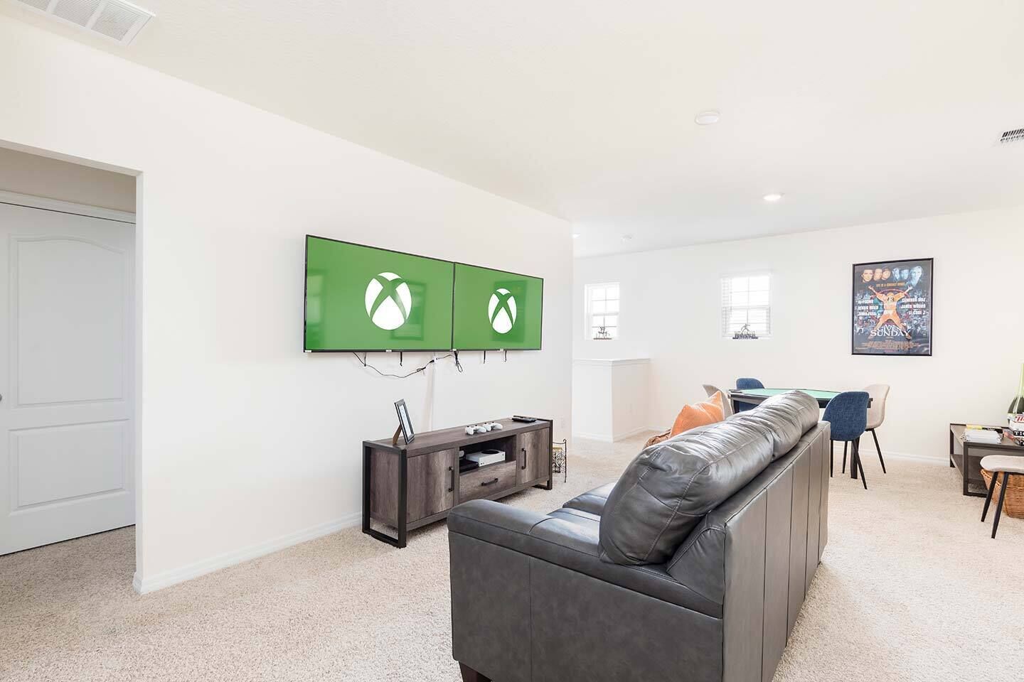 Tilava olohuone, jossa on suuri harmaa nahkasohva, tumma puinen mediakeskus ja seinällä kaksi vihreää Xbox-pelimonitoria.