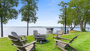 Eine Terrasse am Seeufer mit Adirondack-Stühlen, einem runden Tisch und einer Feuerstelle.