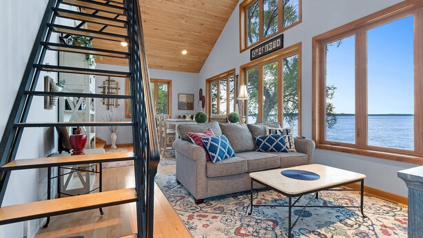 Ein großzügiger Wohnbereich mit einer Holztreppe, einem Sofa mit Zierkissen, einem Couchtisch und großen Fenstern mit Blick auf das Wasser.
