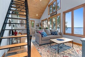 A spacious living area with a wooden staircase, a sofa with decorative pillows, a coffee table, and large windows offering a view of the water. - Cabin Getaway on Lake Noquebay (Crivitz)