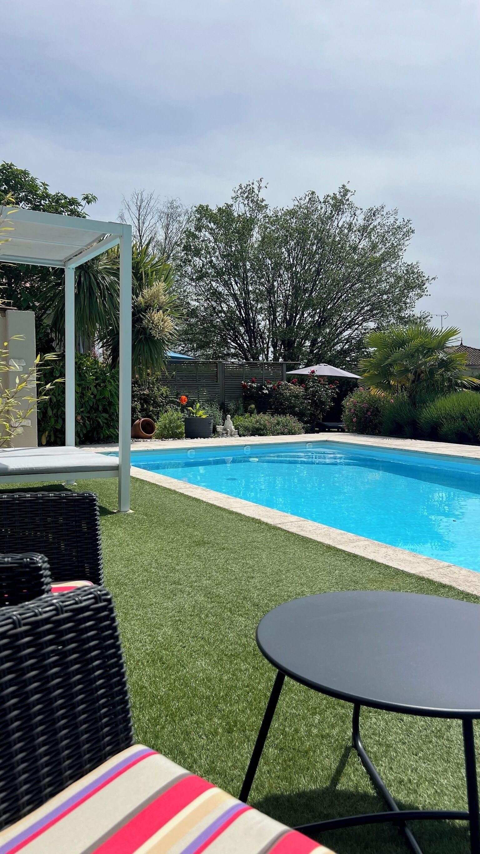 A swimming pool with artificial grass, a white pergola, and wicker furniture.