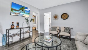 Un salon avec une table basse en verre, un canapé, un téléviseur fixé au mur et un tableau représentant une scène de plage.