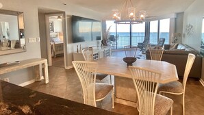 A spacious dining area with a large table and chairs, a view of the ocean through large windows, and a modern kitchen in the background. - Direct Luxury Ocean Front 
Includes umbrella - 2 chairs set up daily 5/31-9/5 (Murrells Inlet)