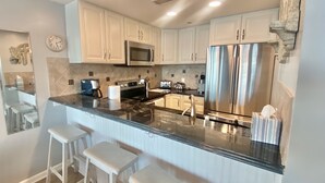 A kitchen with white cabinets, stainless steel appliances, a marble countertop, and bar stools. - Direct Luxury Ocean Front 
Includes umbrella - 2 chairs set up daily 5/31-9/5 (Murrells Inlet)