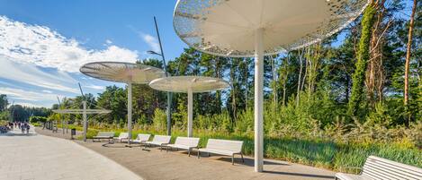 A park with white benches and large decorative umbrellas, surrounded by trees and greenery.