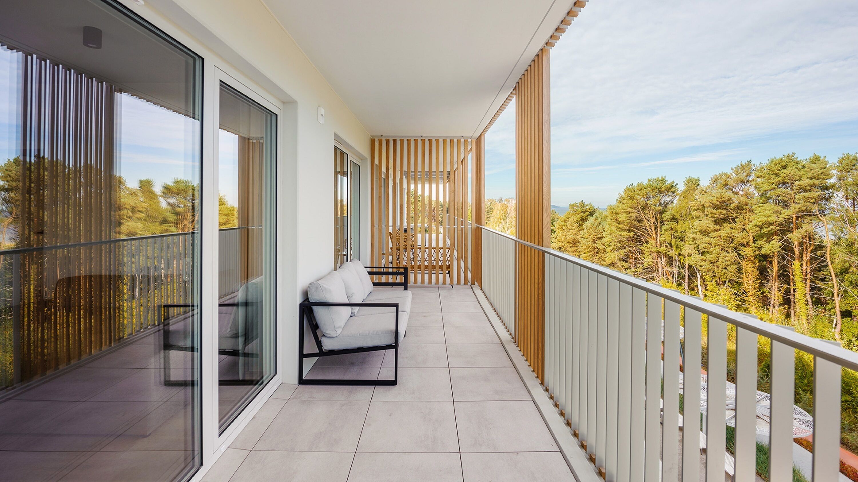 Ein Balkon mit einer Couch und einem Tisch, großen Glasfenstern und Blick auf Bäume.