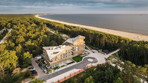 Un immeuble moderne à plusieurs étages, doté d’un jardin paysager à l’avant, situé près d’une plage et entouré d’une forêt dense.