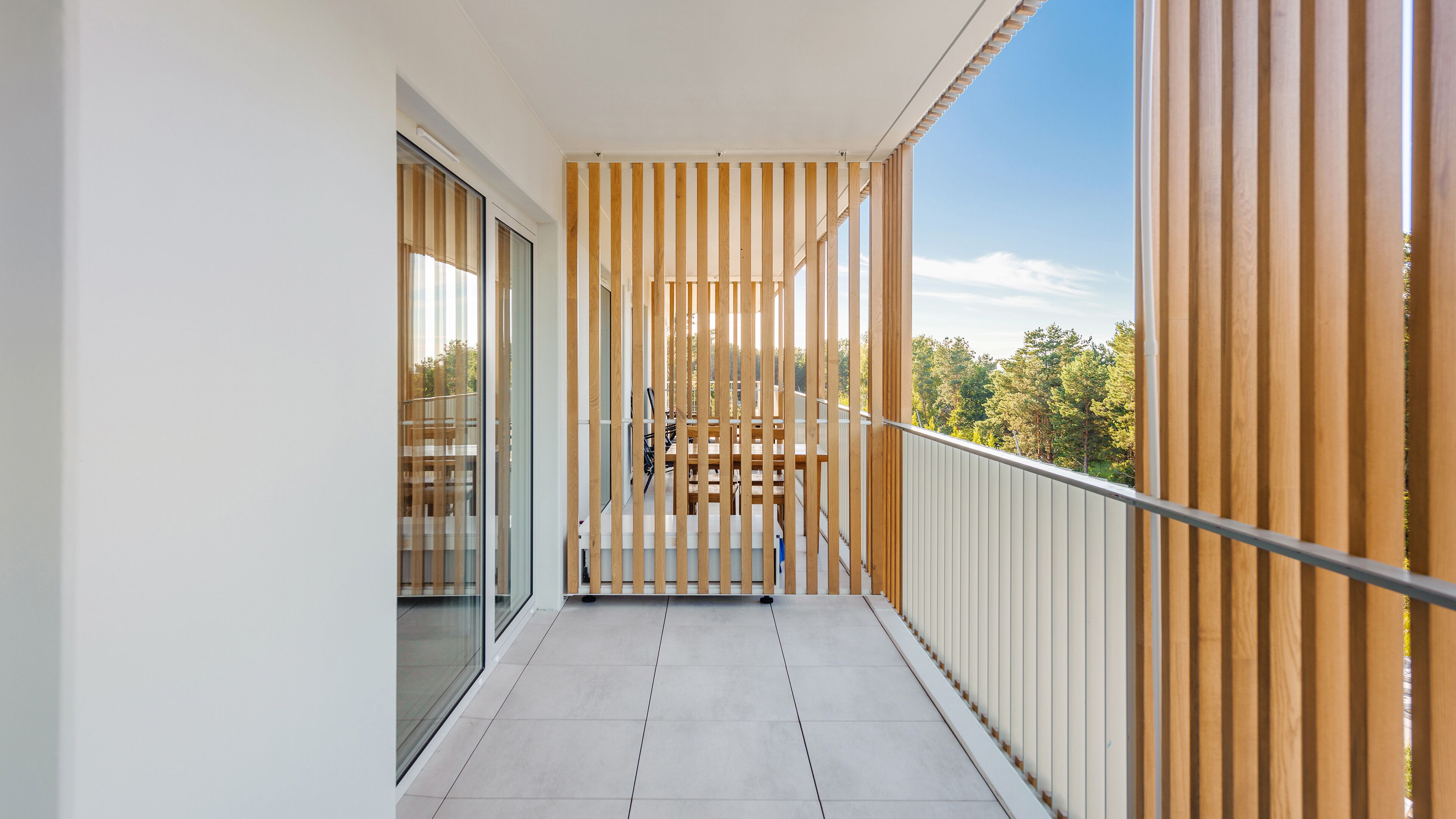 Un balcón estrecho con baranda de madera, puerta de cristal y vistas a árboles y un edificio al fondo.