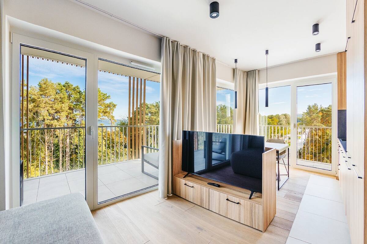 A modern living room with a large TV, sliding glass doors to a balcony, and a view of trees.