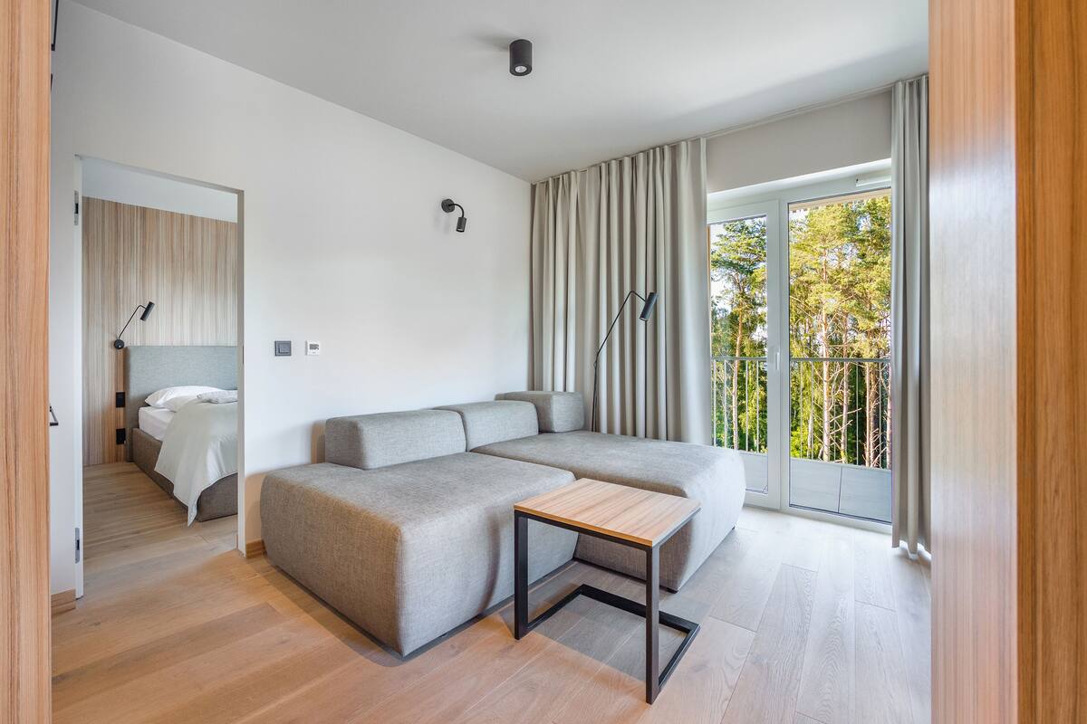 A modern, well-lit bedroom with a large bed, a grey sofa, and a small wooden table.