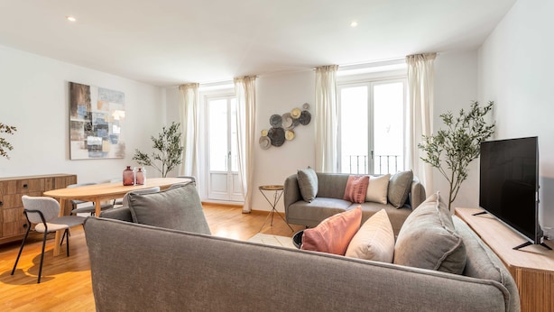 Una sala moderna con un sofá gris, una mesa de comedor de madera y un televisor sobre un mueble de madera.