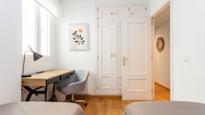Una habitación con escritorio, silla, dos camas, armario y una obra de arte en la pared.
