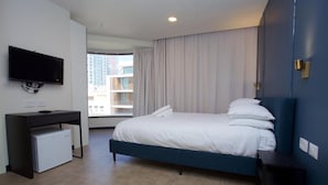 Une chambre d’hôtel avec un lit, un bureau équipé d’un ordinateur, un téléviseur fixé au mur et une fenêtre donnant sur des immeubles.