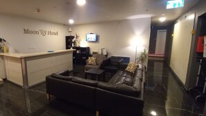 Un espace d’accueil d’hôtel comprenant un comptoir de réception, un canapé, un téléviseur et un mur sur lequel est inscrit le nom de l’hôtel.