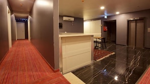 Un couloir d’hôtel avec un tapis rouge, un comptoir de réception, un petit coin salon et une porte donnant sur une chambre.