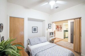 A bedroom with a bed, a ceiling fan, a door, and a hallway leading to a kitchen. - CW C109 River Bend (New Braunfels)