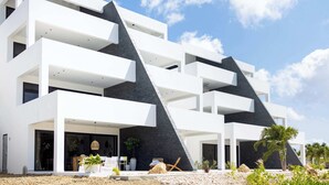A modern multi-story building with balconies, large windows, and a landscaped outdoor area with plants and rocks. - Blue Bay | The Reef | Terrace (Sint Michiel)
