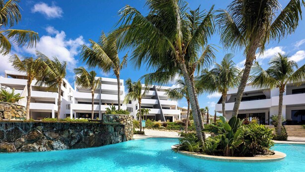 A resort pool with palm trees, a stone wall, and a clear blue sky. - Blue Bay | The Reef | Terrace (Sint Michiel)