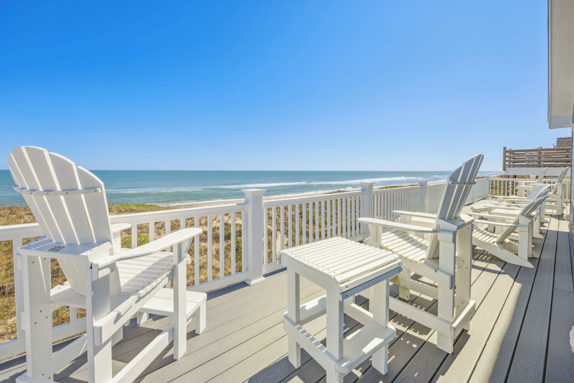 A wooden deck with white Adirondack chairs and a white railing overlooking the ocean.