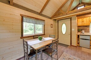 Interior de uma cabana de madeira com mesa de jantar, cadeiras, cozinha e porta com janela circular.