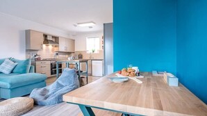 A modern living room with a blue sofa, a wooden dining table with a blue table runner, and a kitchen in the background. - 53 Bibby's Way, Framlingham (Woodbridge)