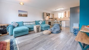 A modern living room with a teal sofa, a small wooden table, and a bean bag chair. A kitchen with white cabinets and a blue wall is visible in the background. - 53 Bibby's Way, Framlingham (Woodbridge)