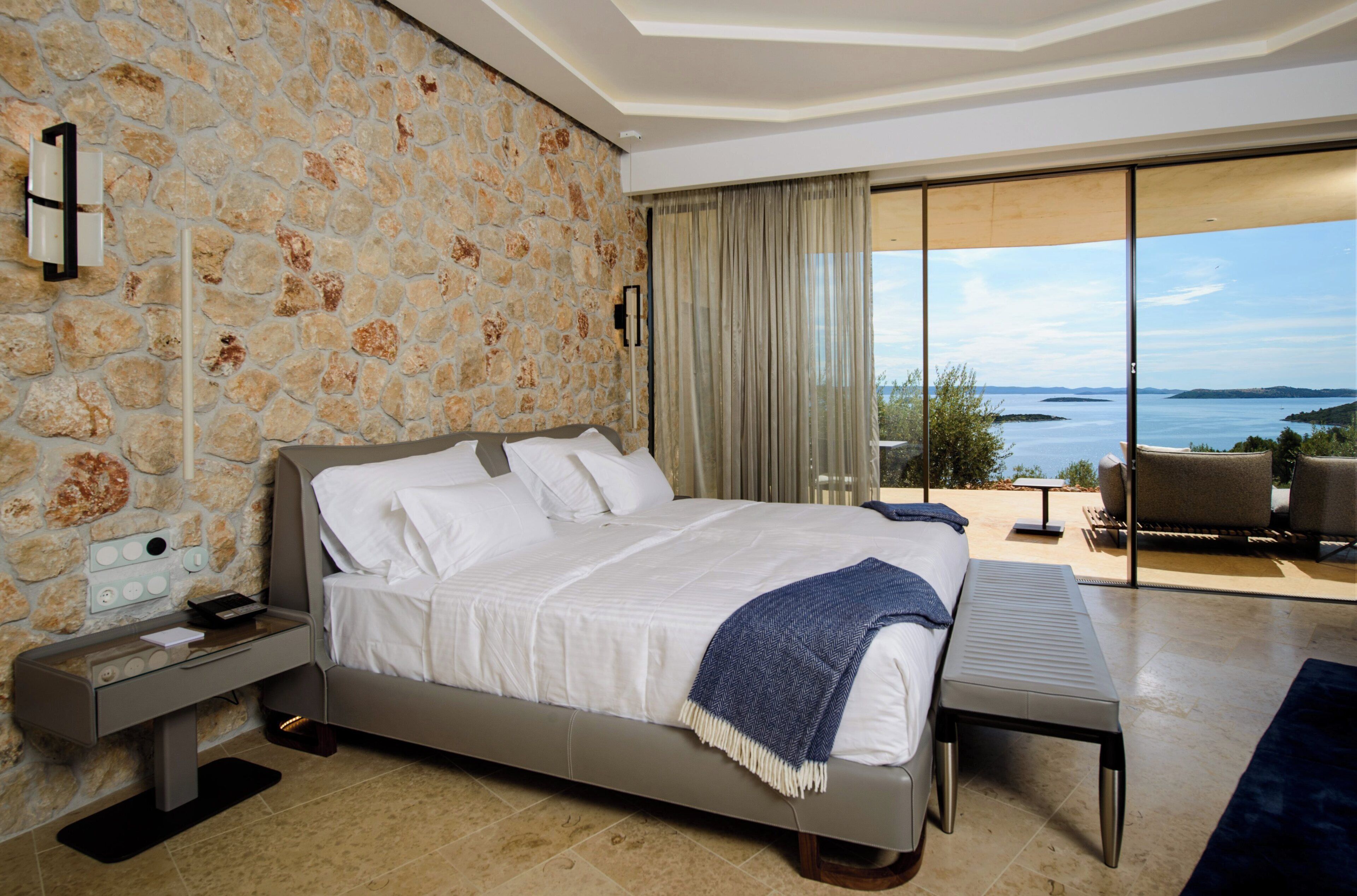 A modern bedroom with a stone wall, a large bed, a bedside table, and a view of the sea through a sliding glass door.