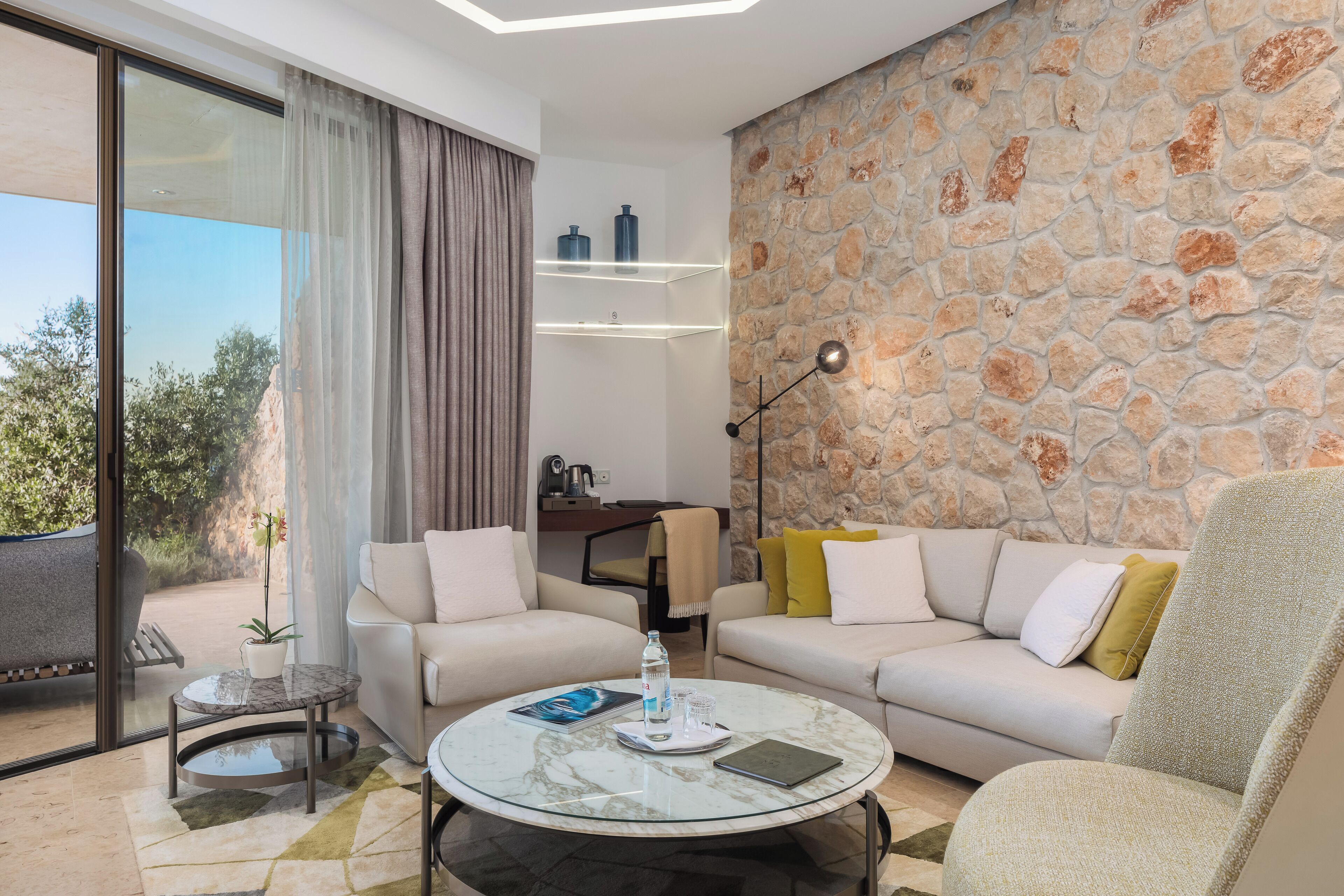 A modern living room with a stone wall, a glass coffee table, two sofas, and a view through a large sliding glass door.
