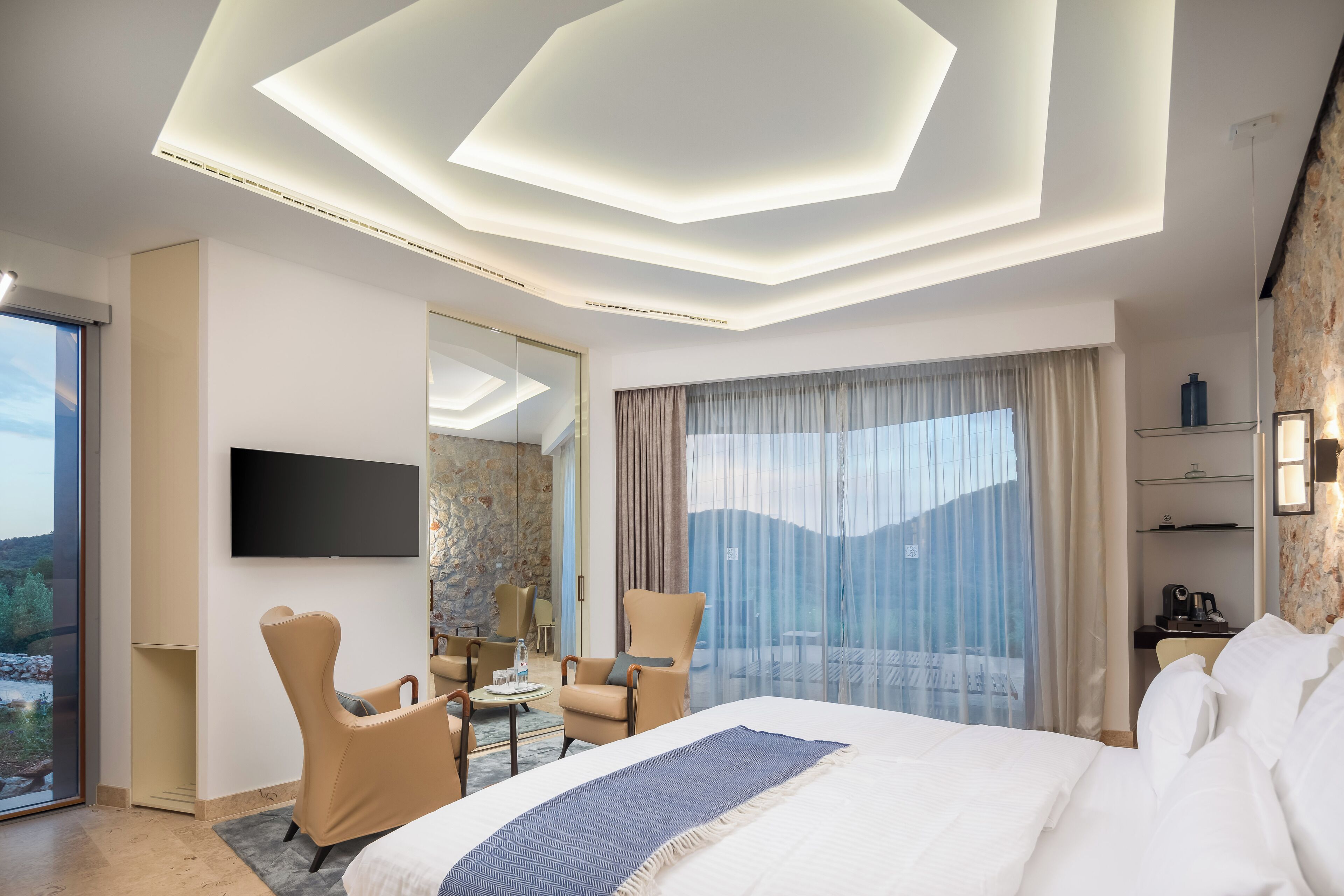 A modern hotel room with a large bed, a flat-screen TV, a seating area with chairs, a dining table, and a balcony with a view.