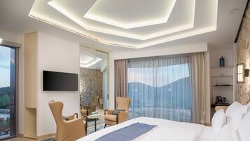 A modern hotel room with a large bed, a flat-screen TV, a seating area with chairs, a dining table, and a balcony with a view.