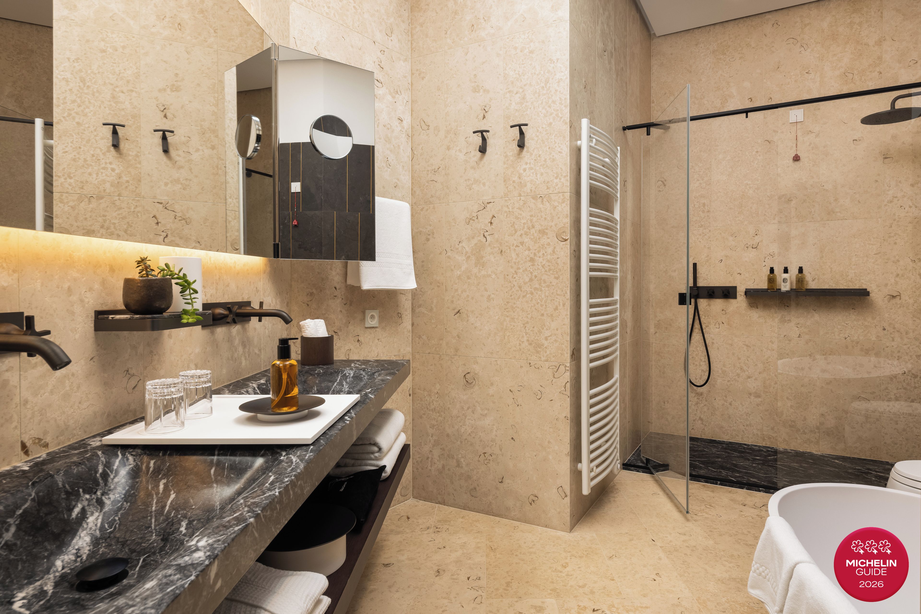 Un baño moderno con encimera de mármol, cabina de ducha de vidrio y un cartel de Guía Michelin 2026.
