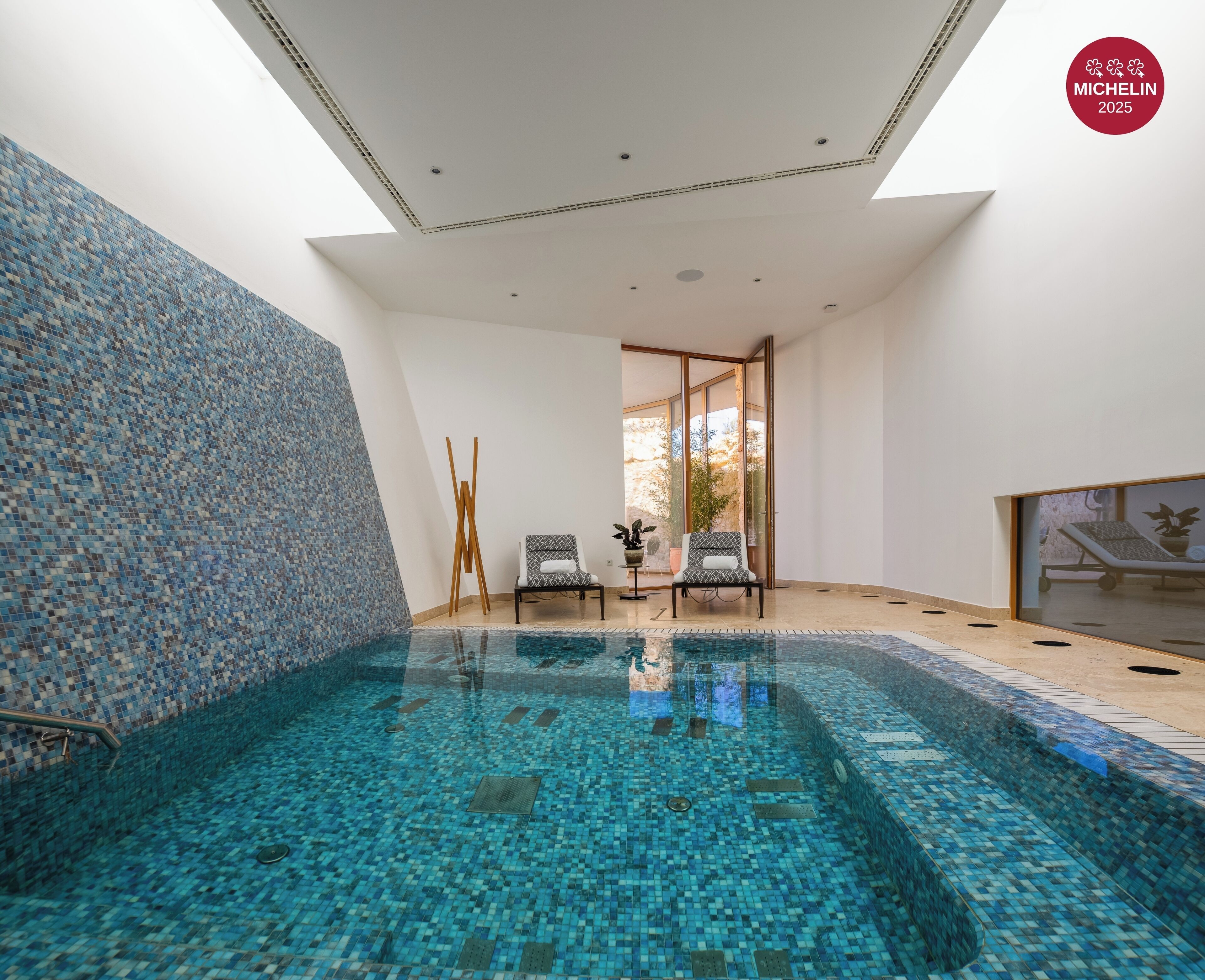 A modern indoor swimming pool with mosaic tile walls and ceiling, two lounge chairs, and a glass door leading outside.