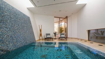 A modern indoor swimming pool with mosaic tile walls and ceiling, two lounge chairs, and a glass door leading outside.
